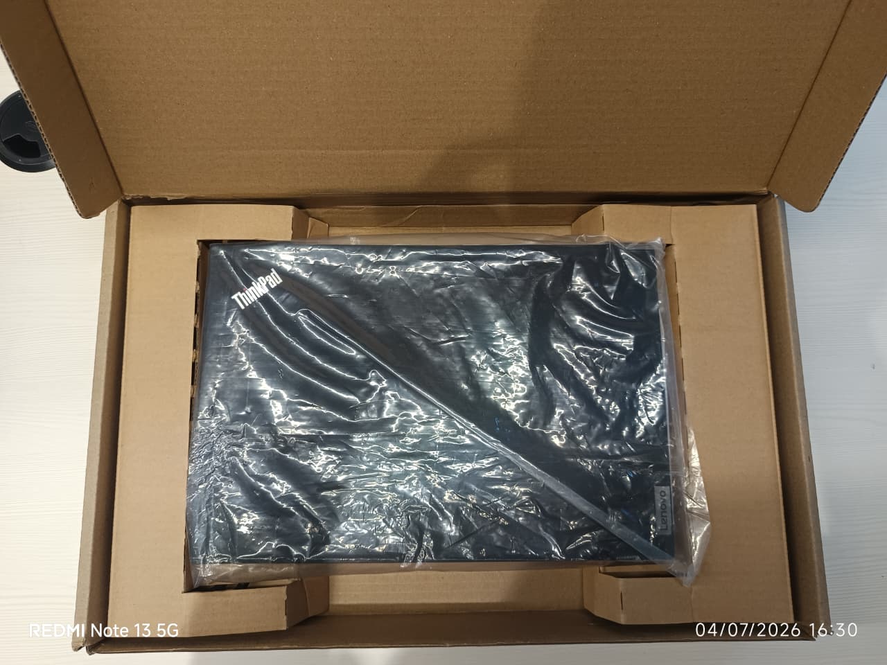 A black Lenovo laptop inside its  brown cardboard packaging box, still wrapped in protective plastic.