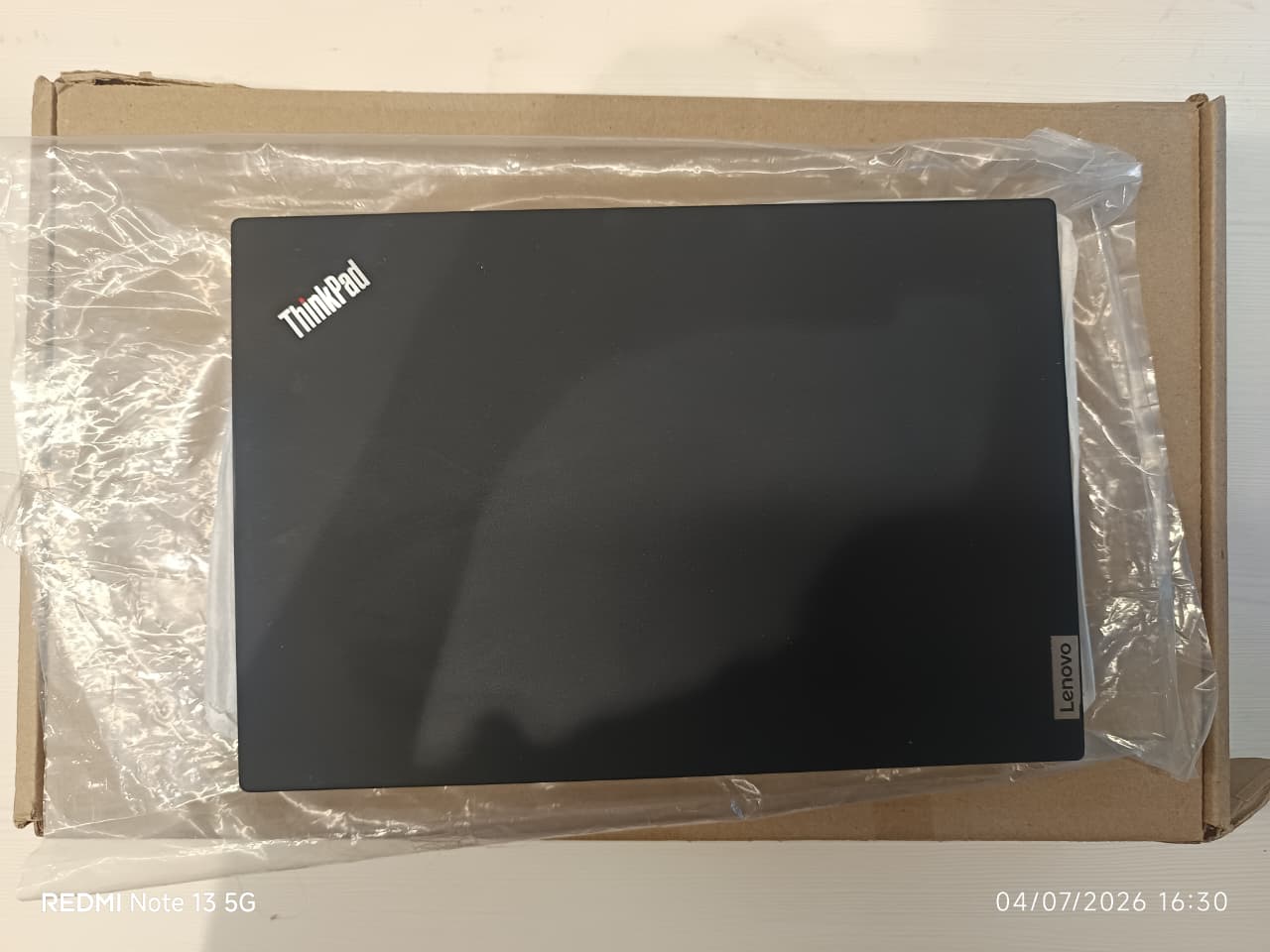 A black Lenovo ThinkPad laptop inside a brown cardboard box, wrapped in clear plastic protective film. The ThinkPad logo is visible in the top-left corner of the laptop lid, and a Lenovo logo badge is visible on the bottom-right corner.