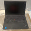 A black Lenovo ThinkPad laptop on a white surface, partially wrapped in protective plastic. The screen is turned off, and the keyboard, trackpad, and red TrackPoint nub are clearly visible.