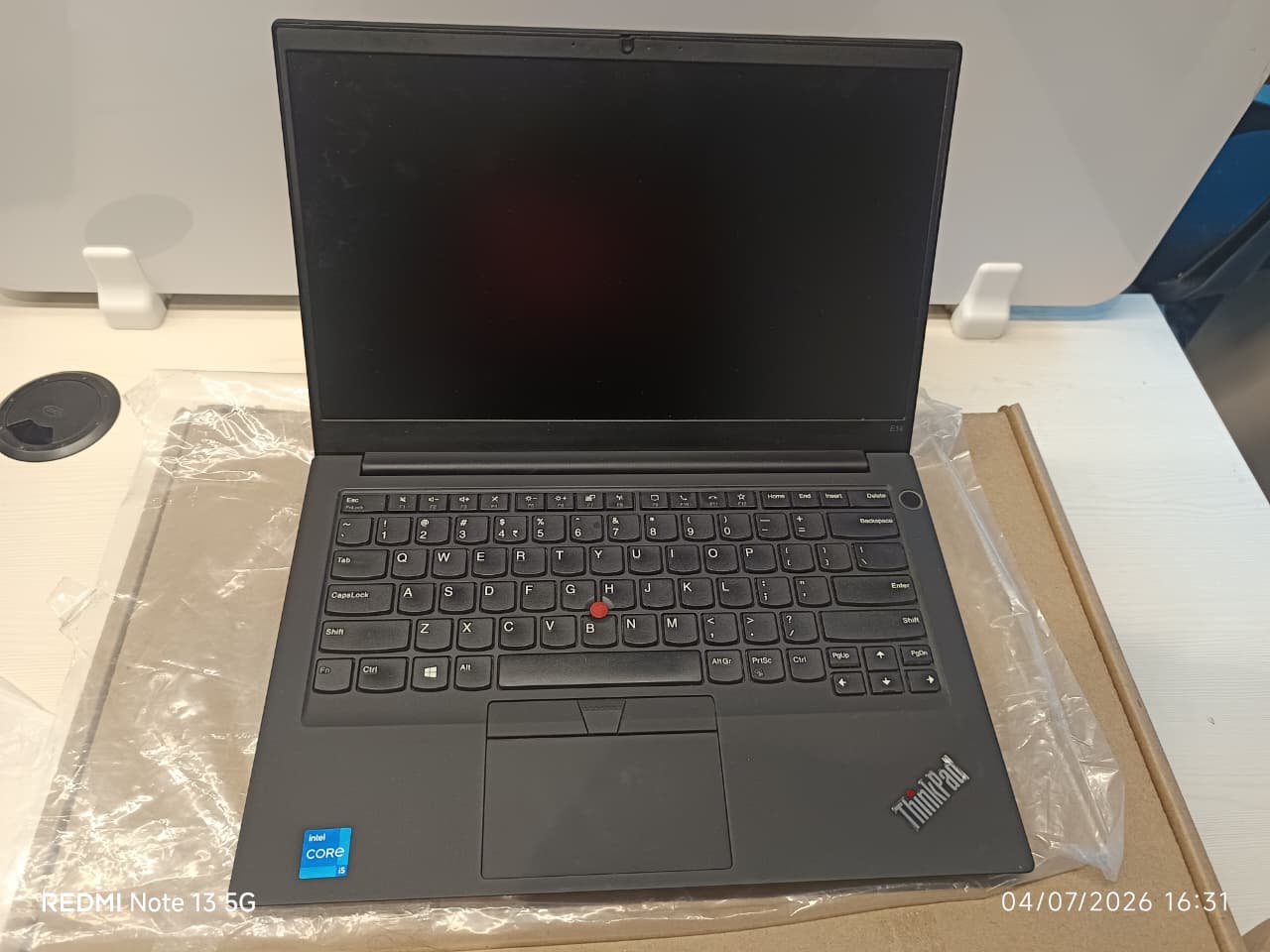 A black Lenovo ThinkPad laptop on a white surface, partially wrapped in protective plastic. The screen is turned off, and the keyboard, trackpad, and red TrackPoint nub are clearly visible.