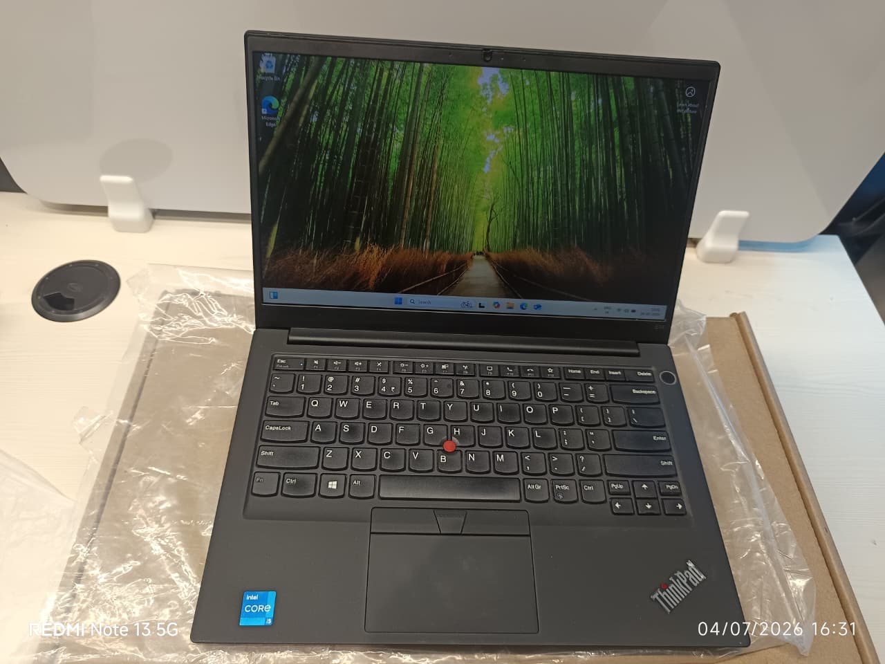 A black Lenovo ThinkPad laptop on a white surface, partially wrapped in protective plastic. The screen is turned on, and the keyboard, trackpad, and red TrackPoint nub are clearly visible.